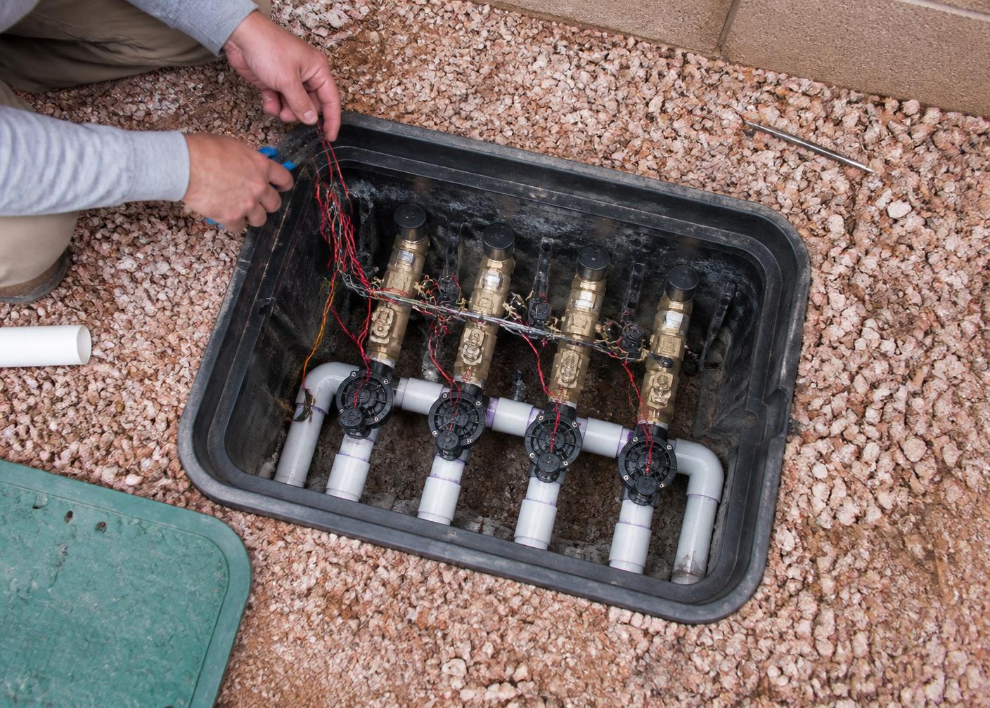Open irrigation valve box showing anti-siphon valves, a PVC manifold and control wiring being serviced in an Arizona side yard